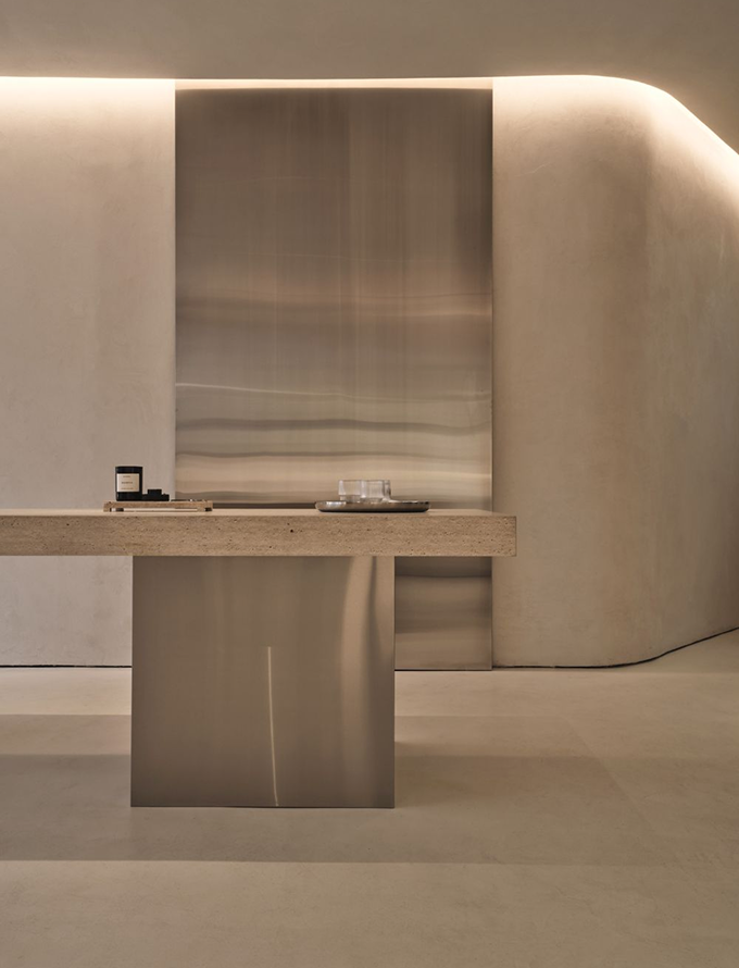 Modern minimalist interior with textured concrete walls, a large stainless steel panel, and a beige stone table with a metal base.