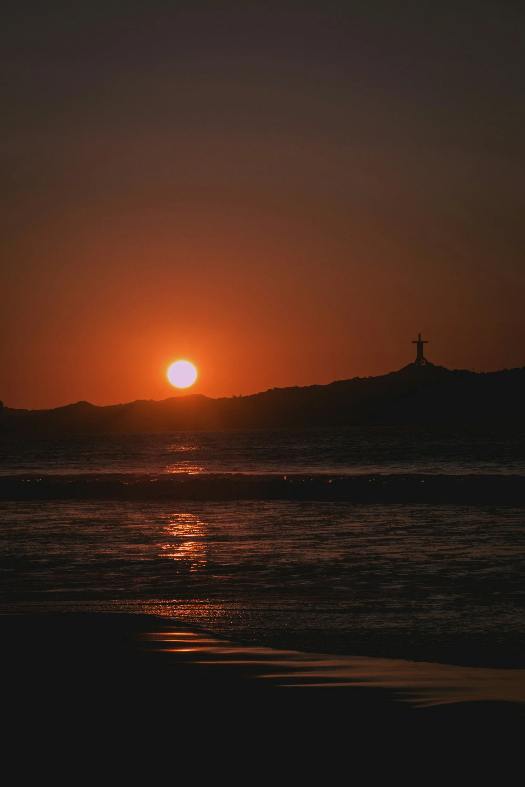 Atardecer en la playa con el sol en el horizonte y una cruz en la cima de una colina.