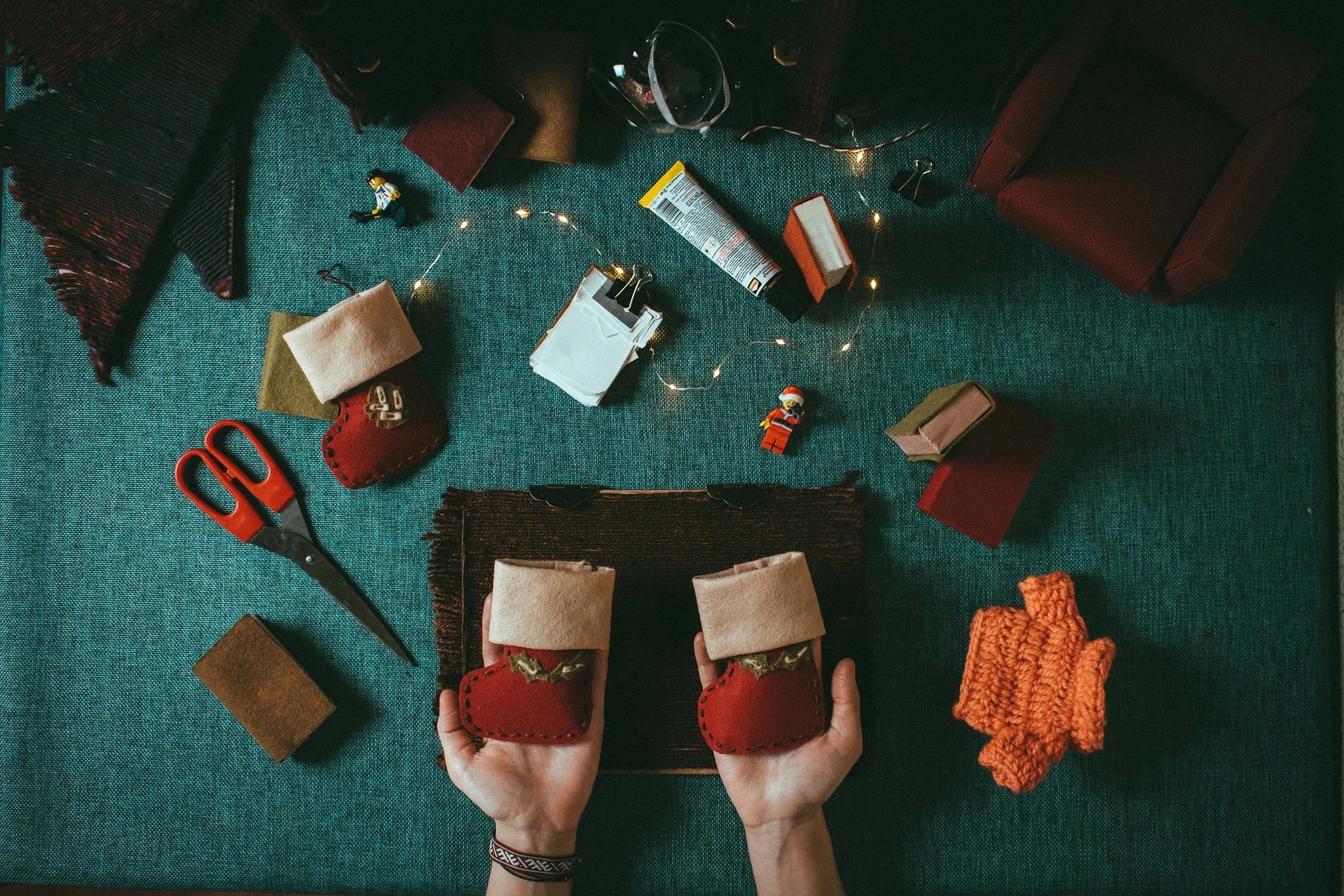 Persona sosteniendo una casa de fieltro roja, rodeada de materiales para hacer manualidades, incluyendo tijeras, botones, fieltro, bobinas de hilo, figuritas decorativas y otros objetos decorativos en una mesa con tapete verde