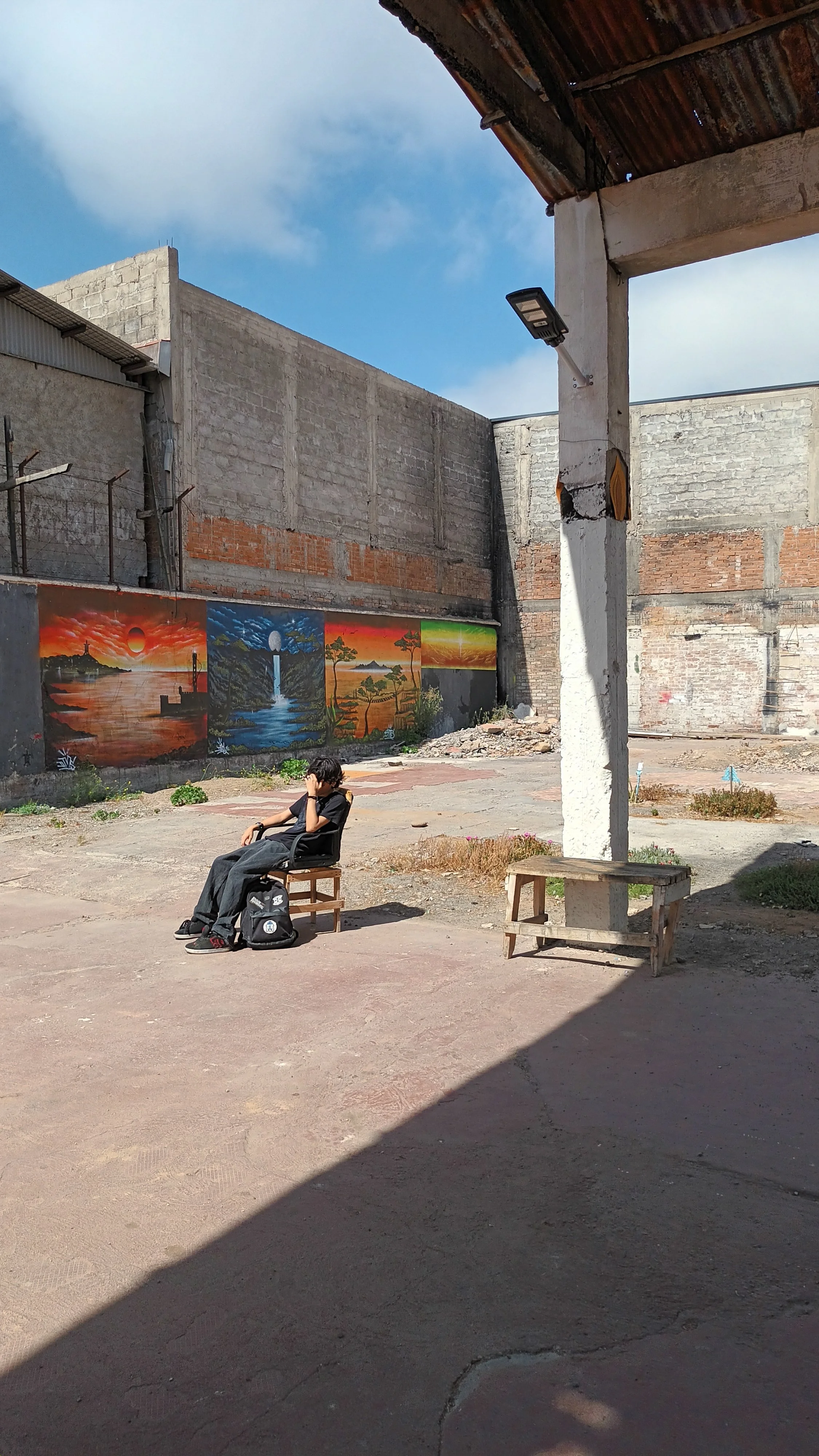 Un espacio abierto con un muro decorado con pinturas de paisajes, una persona sentada en una banca y un poste con un foco de luz, en un día soleado con cielo despejado
