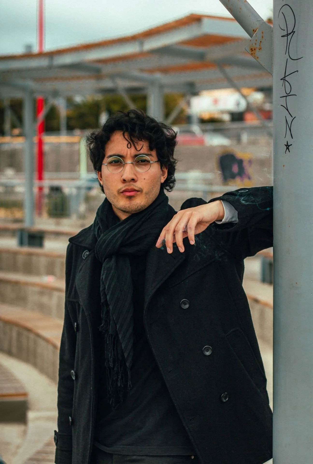 Un hombre de cabello rizado y gafas redondas posa apoyado en una baranda de metal en un parque o espacio abierto, vestimenta oscura y expresión seria.
