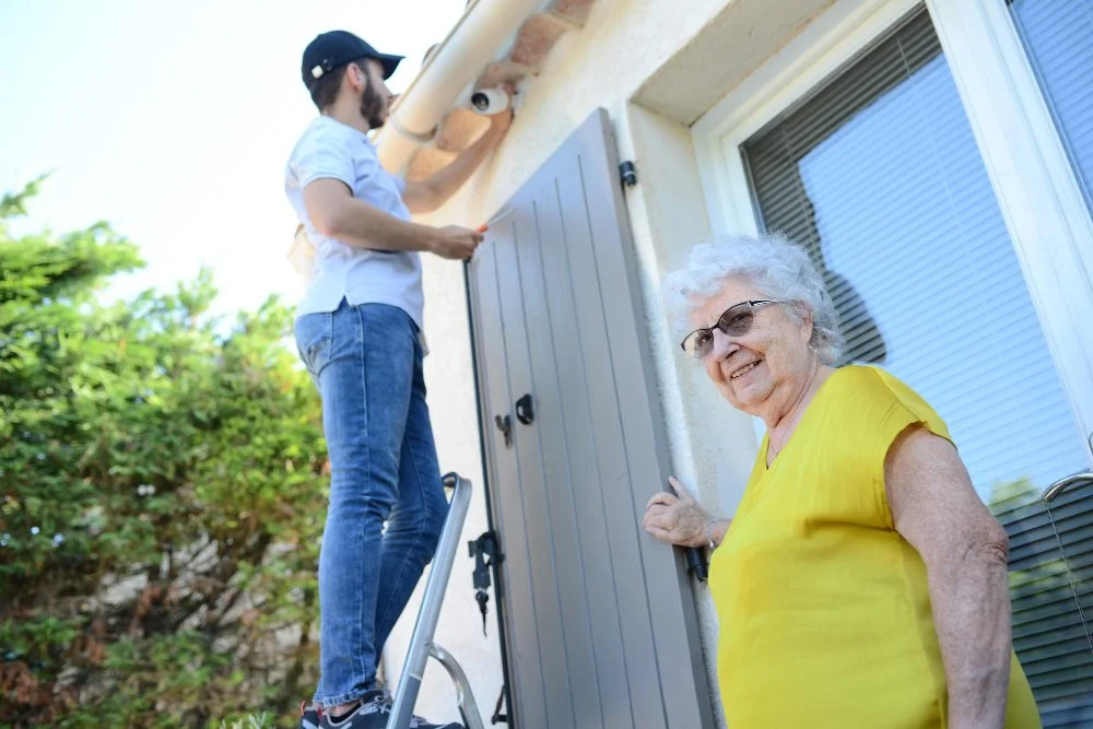 Young man repairing or installing a roof or gutter while standing on a ladder outside a house, with an elderly woman smiling and watching from the doorway.