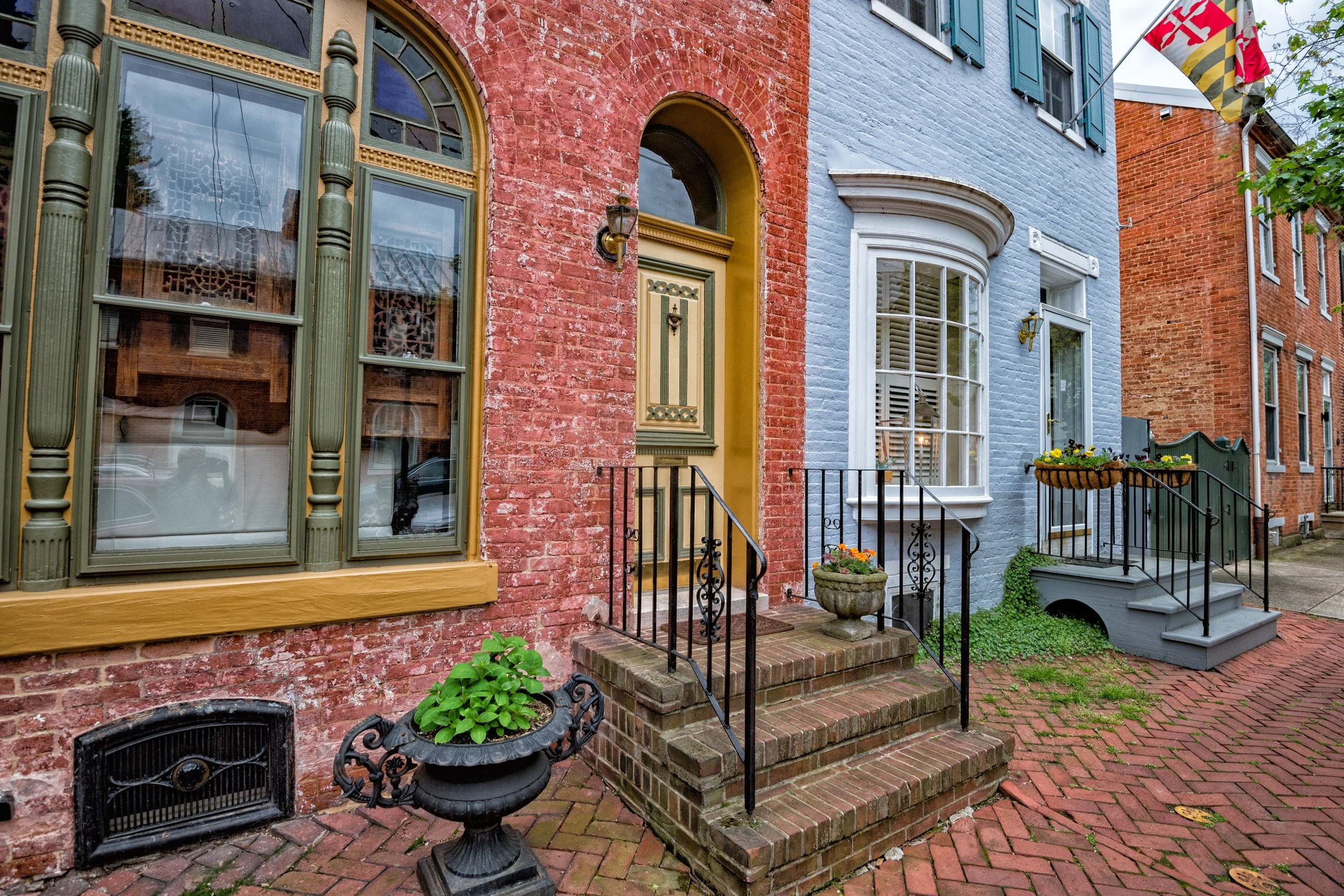 A row of colorful brick townhouses with front steps, decorative window trim, and flower planters on a brick sidewalk.