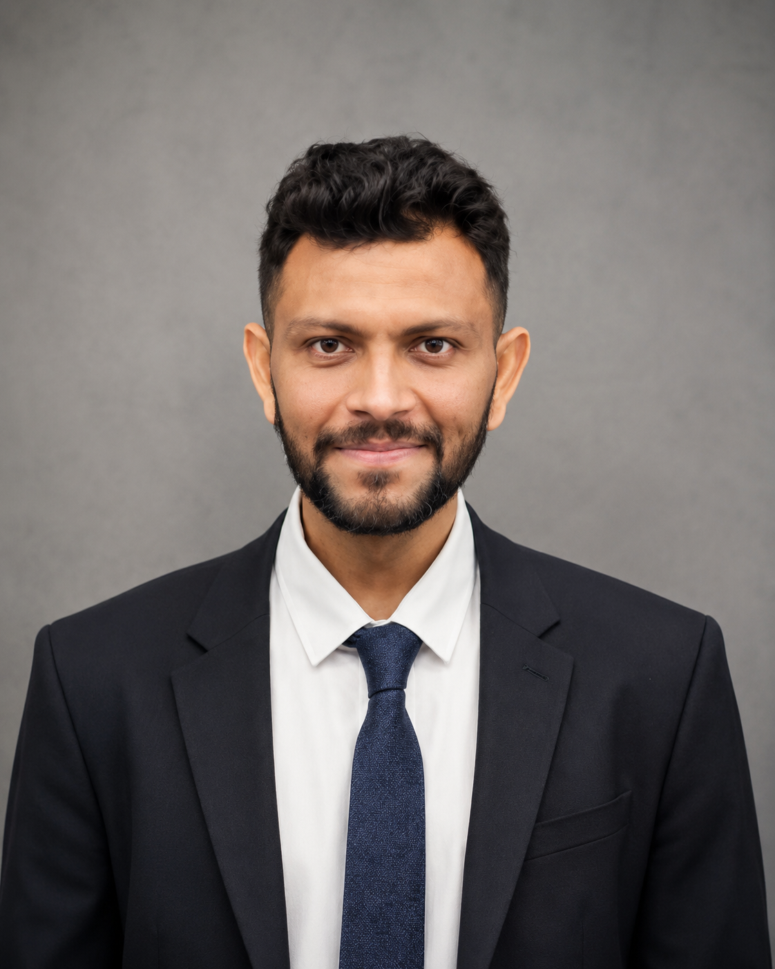 Portrait of a young man with dark hair, beard, in a black suit, white shirt, and blue tie, smiling against a gray background.