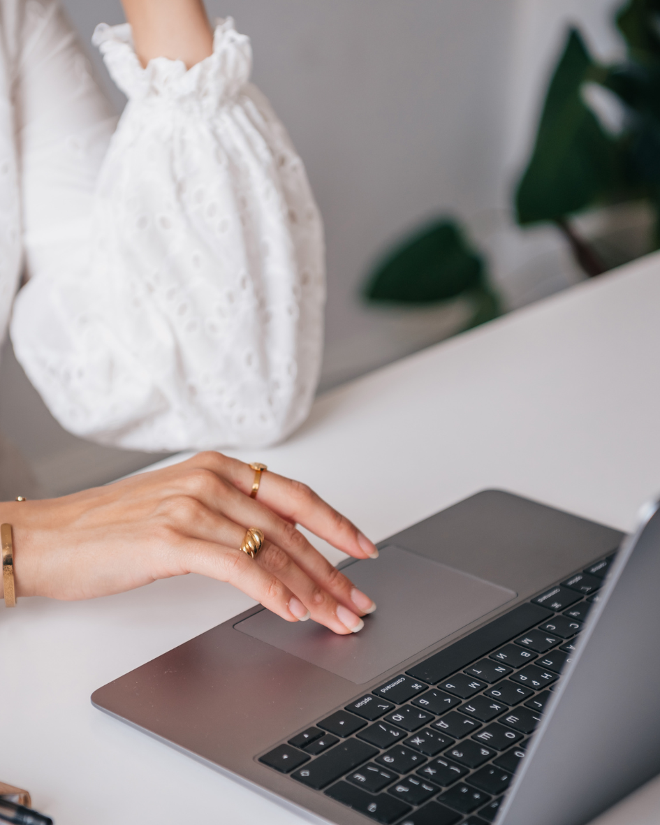 Person using a laptop with their hand on the trackpad, wearing jewelry, in front of a white lace garment and some green leaves in the background.