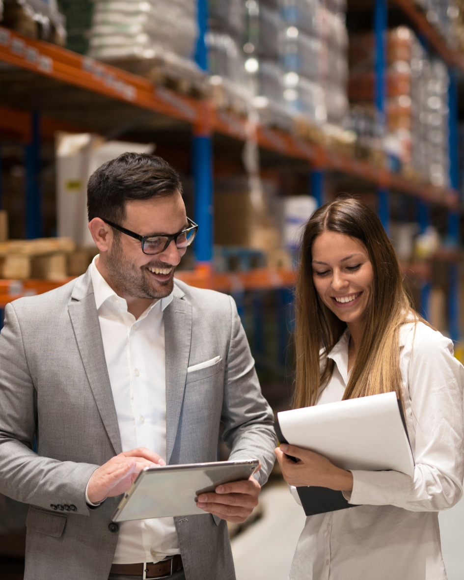 Two people, a man in a gray suit and glasses and a woman with long brown hair in a white shirt, stand in a warehouse talking and looking at a tablet and a notebook.