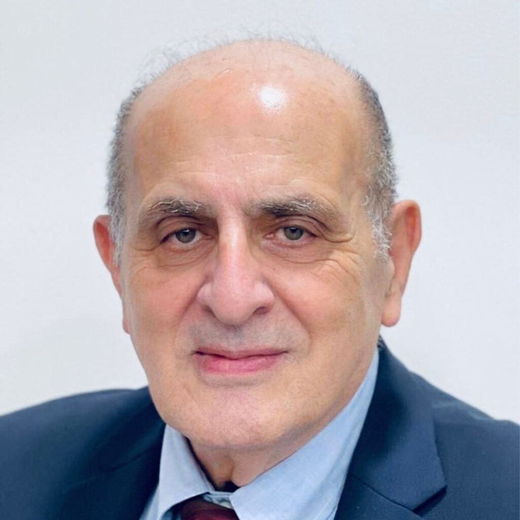 Headshot of an older man with a bald head, light skin, and blue eyes, wearing a dark suit and tie, against a plain light background.