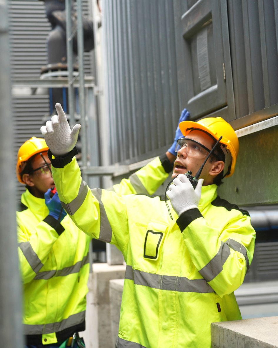 Two workers in high-visibility yellow jackets and yellow hard hats, working outdoors near industrial equipment. One worker is speaking into a radio, gesturing upward, while the other is standing nearby.