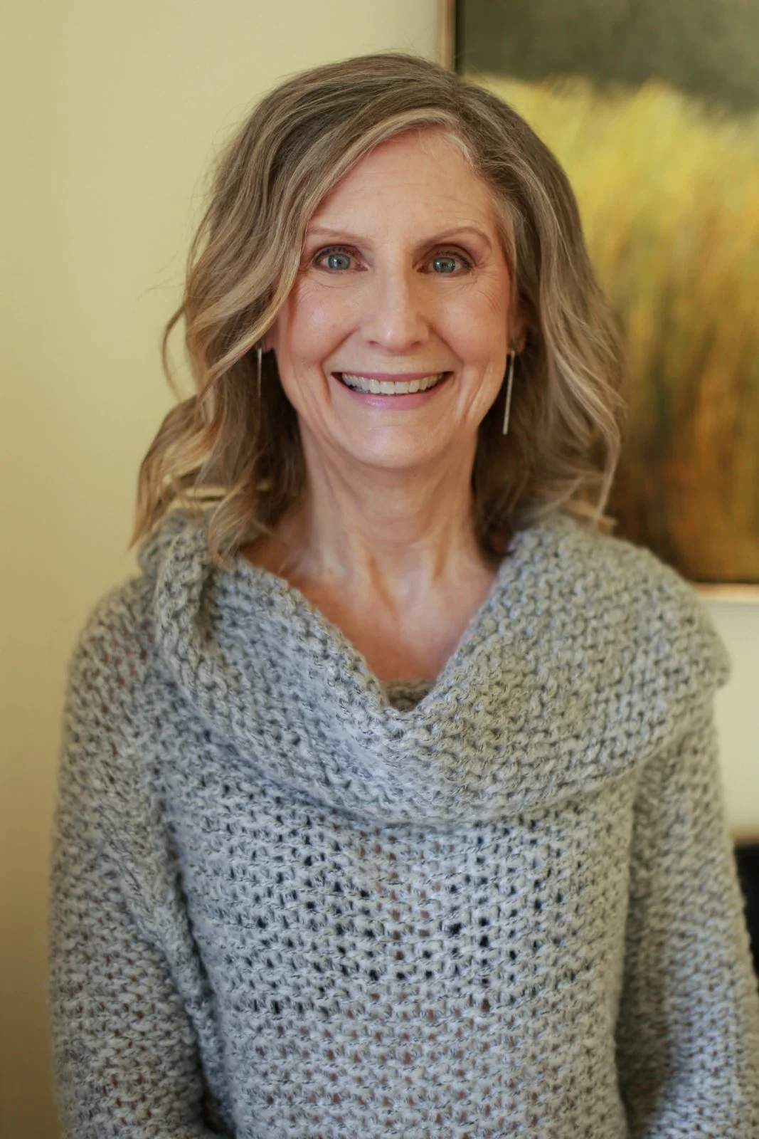 A smiling woman with shoulder-length wavy hair, wearing a chunky knit sweater, standing indoors with a blurred natural artwork in the background.