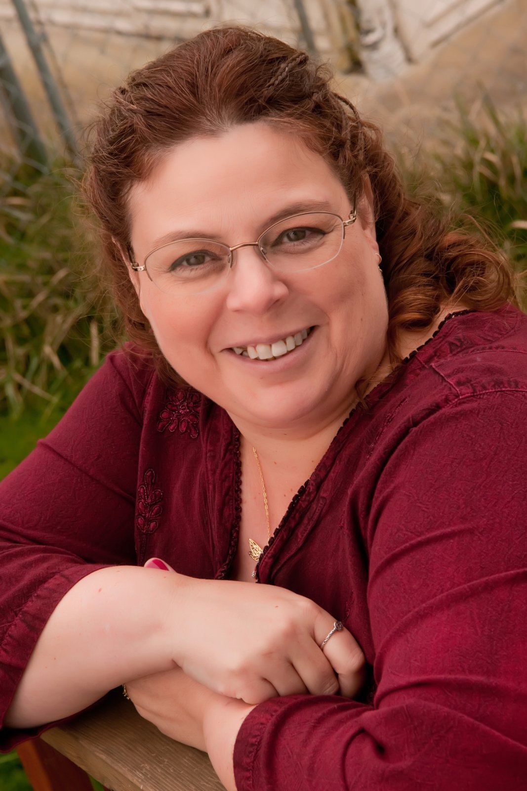A woman with red hair, glasses, and a maroon top smiling outdoors with greenery and garden plants in the background.