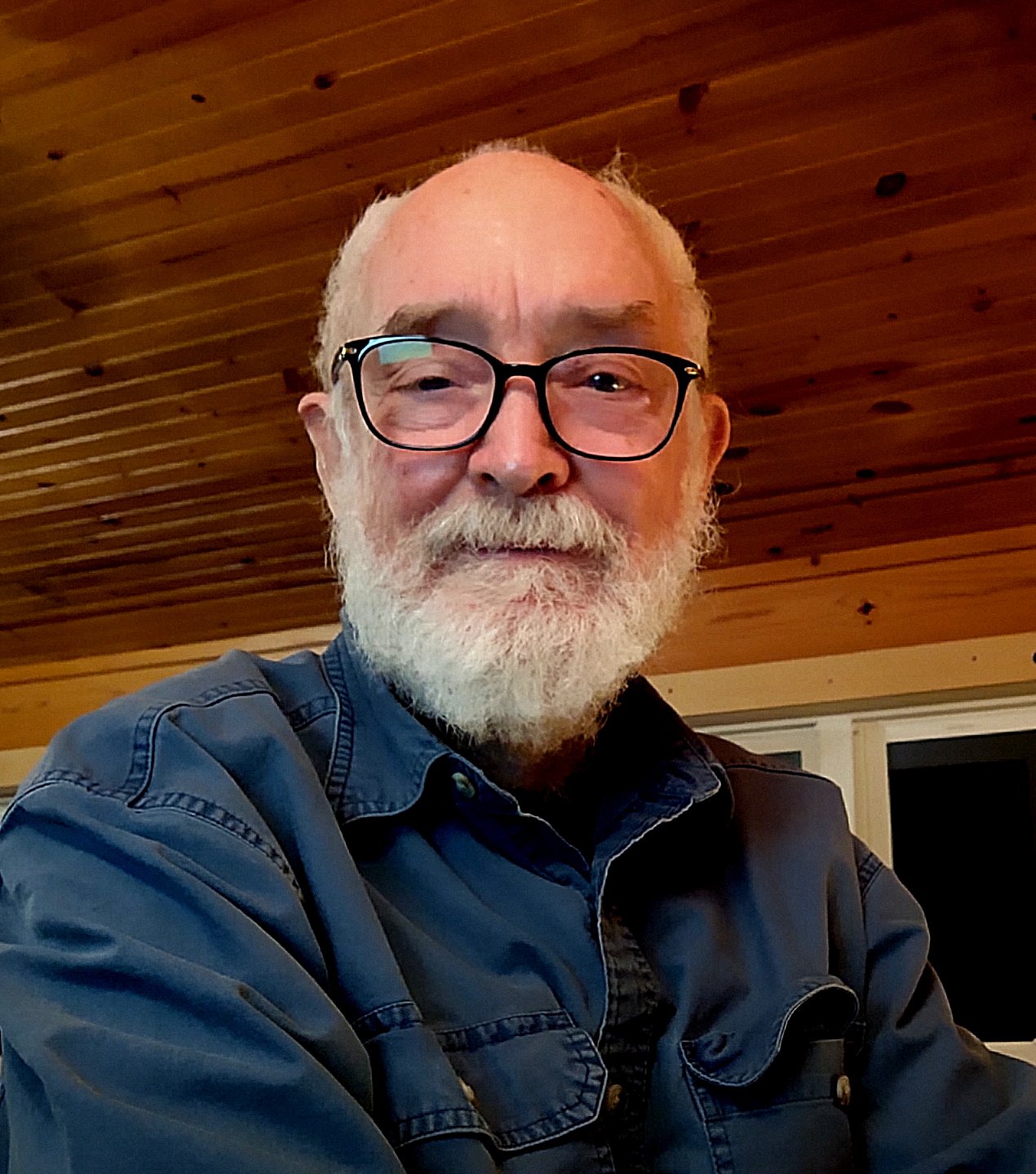 An elderly man with a white beard, glasses, and a blue denim jacket smiling at the camera in a wooden ceiling room.