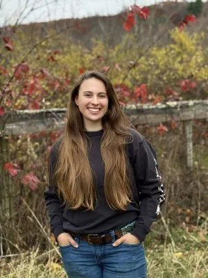 Young woman with long brown hair smiling outdoors in front of autumn trees and a wooden fence.