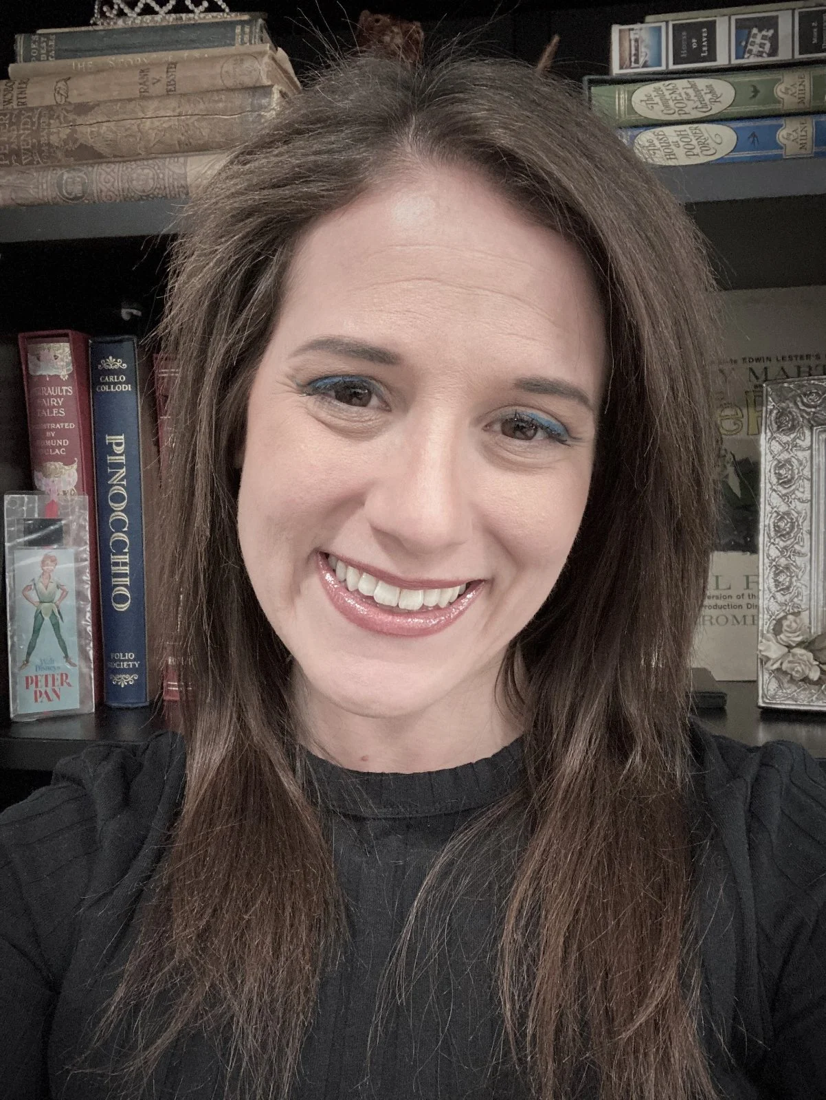 Close-up of a woman with long brown hair, smiling, standing in front of a bookshelf filled with books and decorative items.
