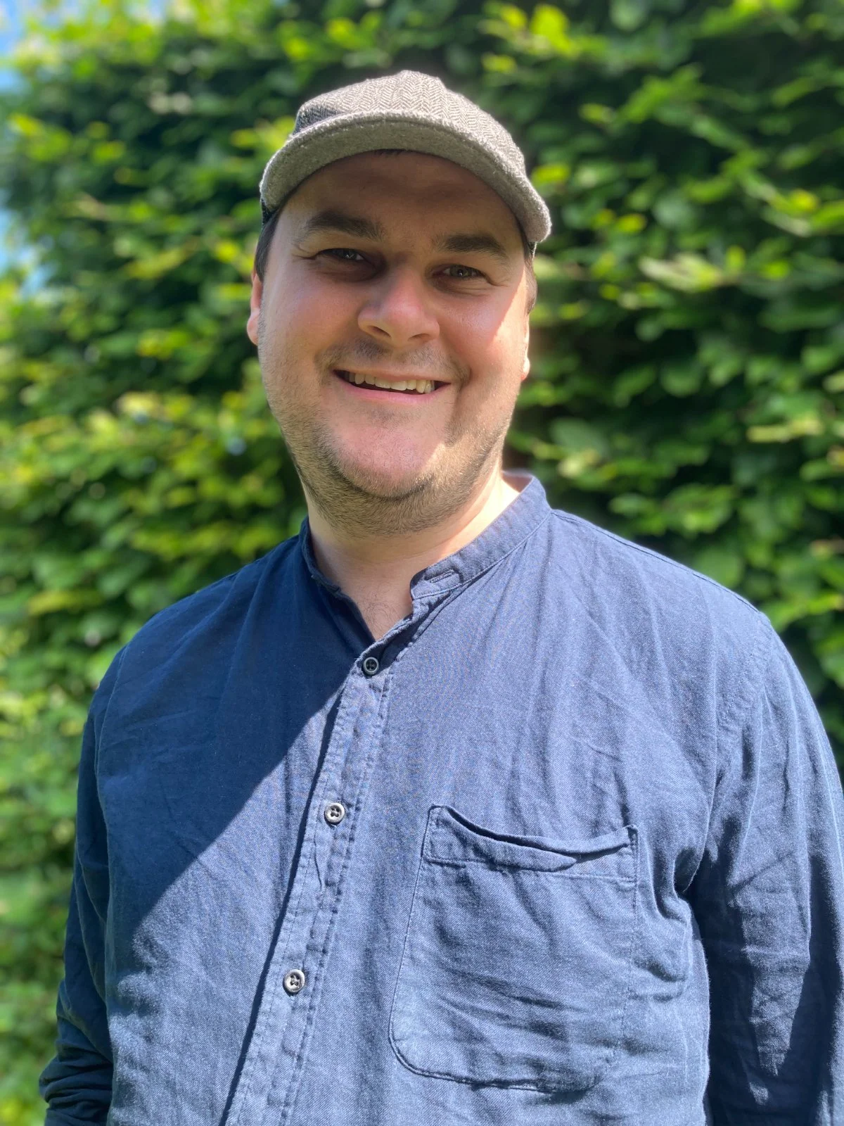 A smiling man wearing a gray cap and a dark blue button-up shirt, standing outdoors in front of green foliage.
