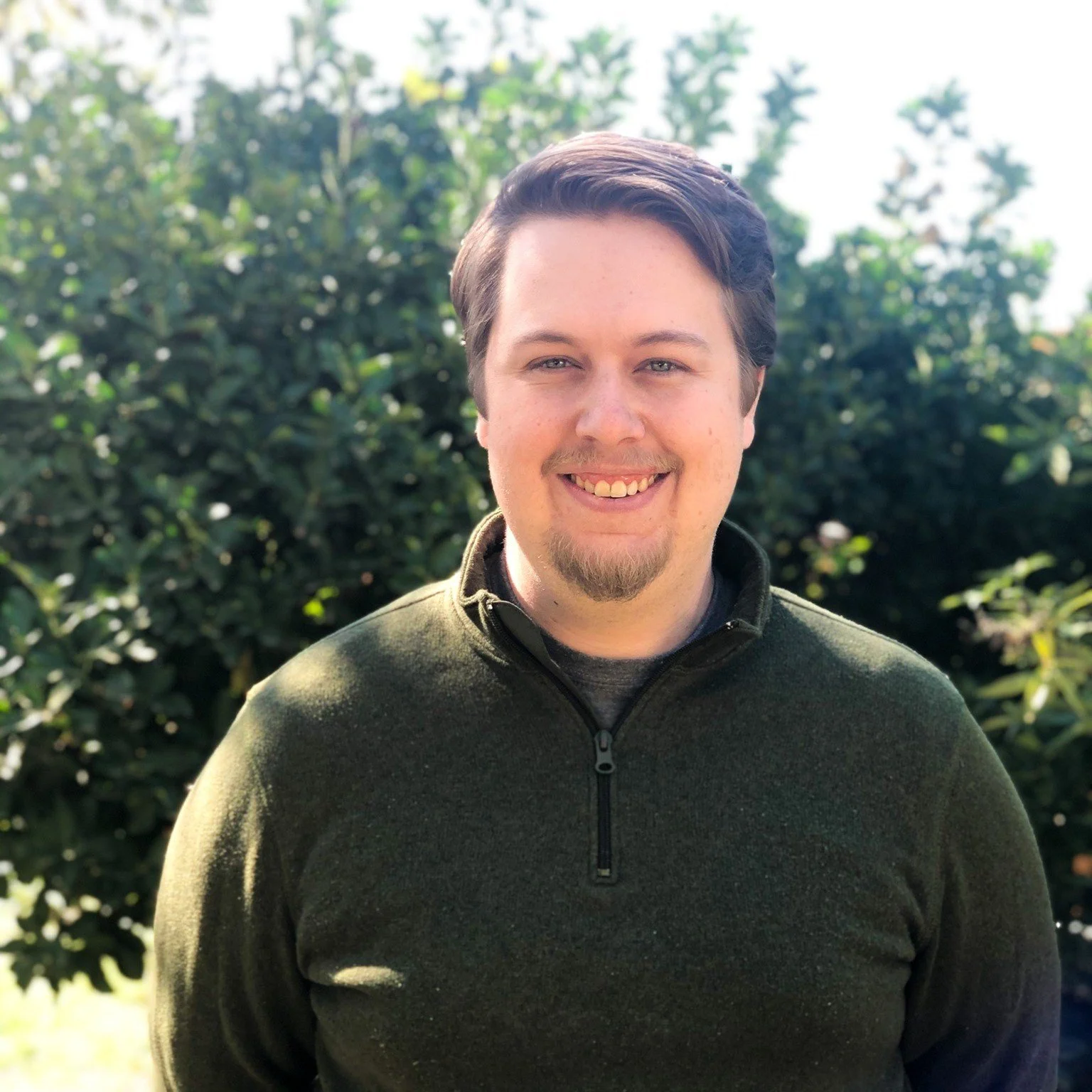 A smiling young man with short dark hair and a beard, wearing a green quarter-zip fleece, standing outdoors in front of dense green foliage.