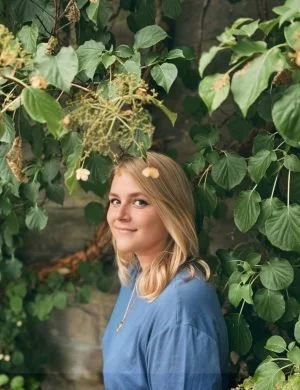 A young woman with blonde hair smiling in front of green leafy plants.