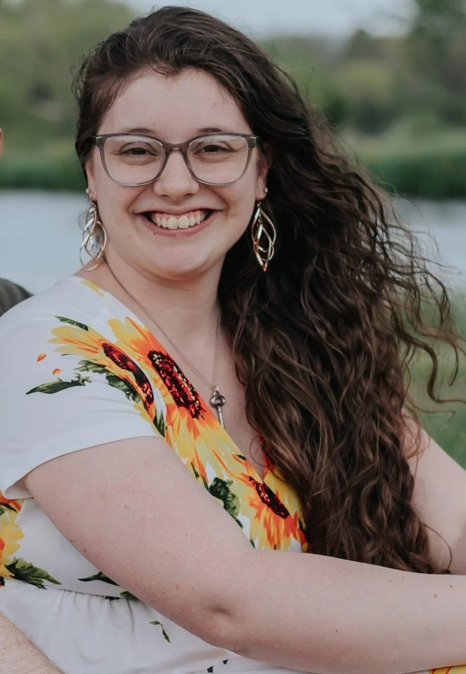 A woman with long curly brown hair, glasses, and earrings, smiling outdoors near a body of water, wearing a white top with a sunflower pattern.