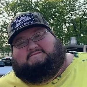 A man with glasses, a beard, and a cap featuring a logo, outdoors with trees in the background.