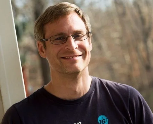 A man with glasses and light brown hair smiling outdoors near a window, with trees in the background.