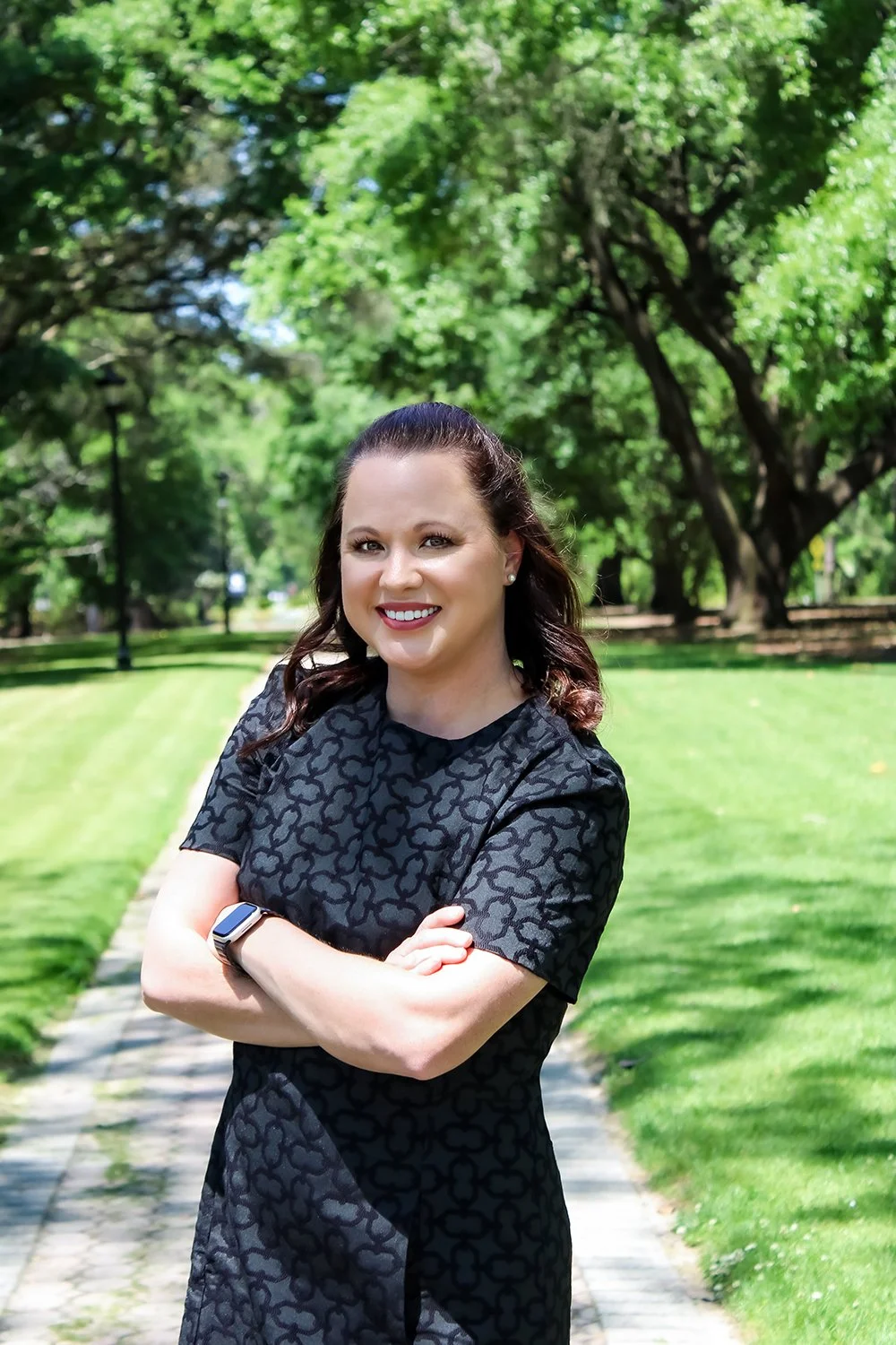 Smiling woman in a black patterned dress with crossed arms standing on a park sidewalk with green grass and trees in the background.