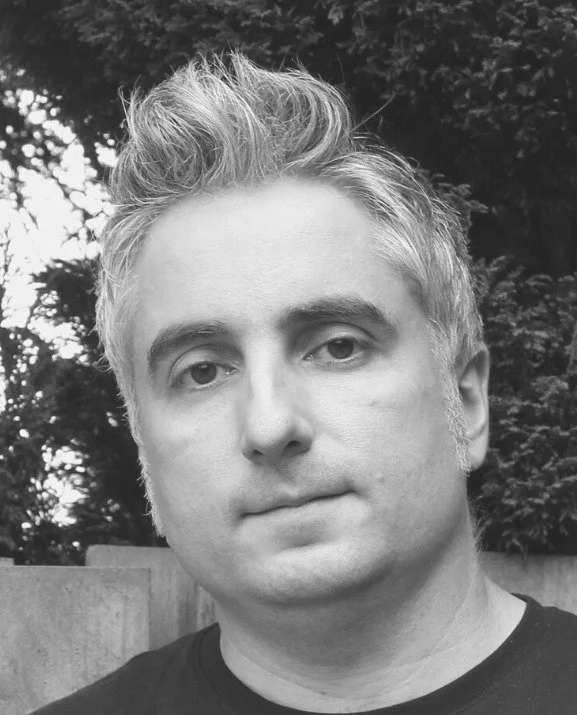 A man with short, spiked gray hair in an outdoor setting, with trees and a fence in the background.
