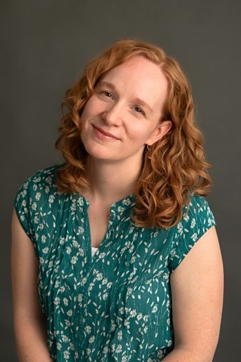 A woman with curly red hair wearing a green patterned blouse, smiling at the camera, against a plain background.