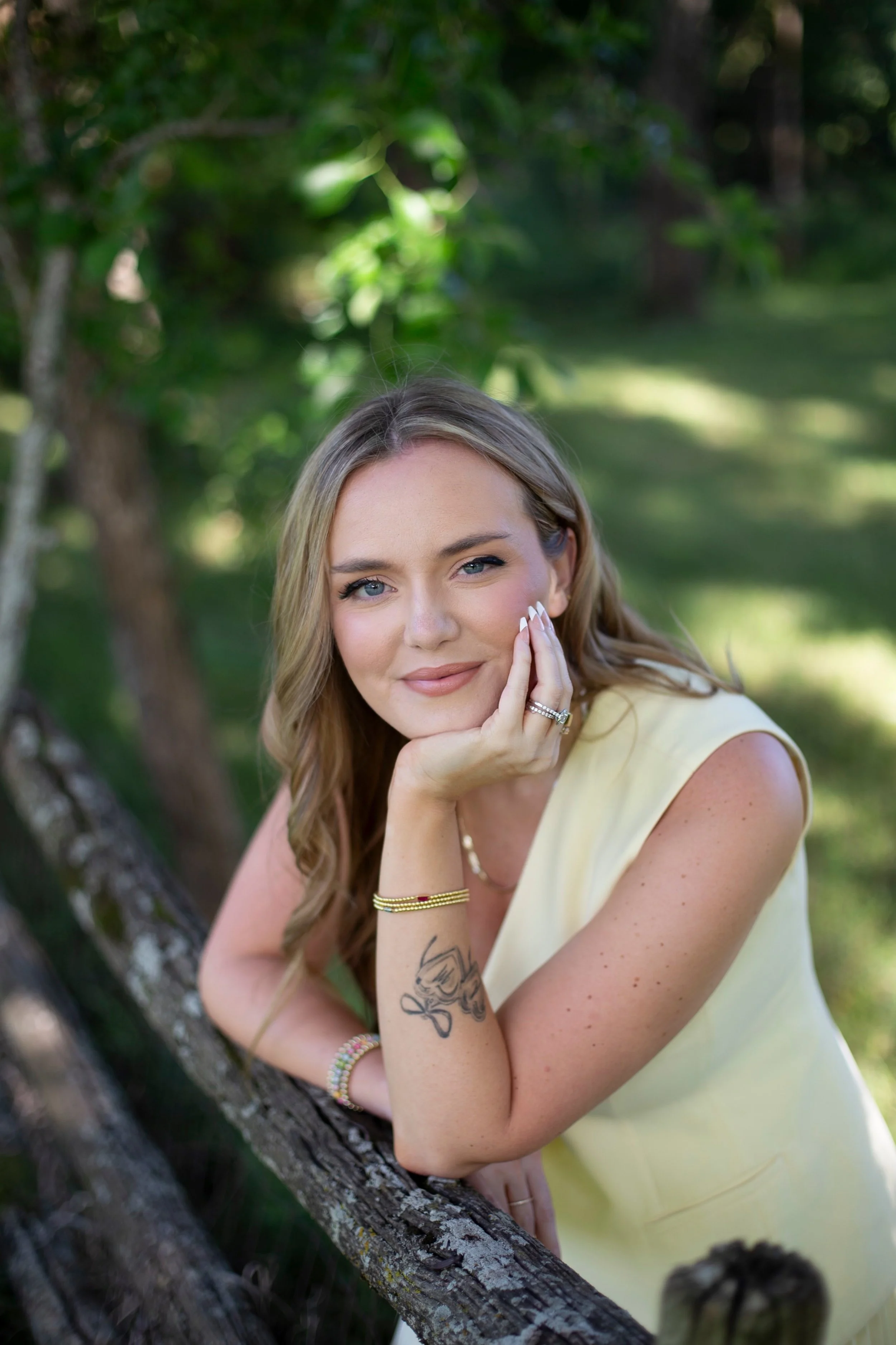 A woman with long light brown hair, fair skin, and blue eyes, leaning on a tree outdoors with green foliage background. She is smiling softly and resting her chin on her hand, wearing a yellow sleeveless top, multiple bracelets, and rings, with a tattoo on her left forearm.