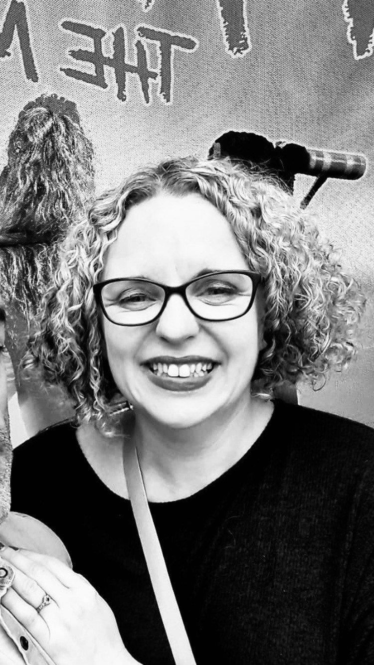 A woman with curly hair wearing glasses and a black shirt, smiling for a photo. She has rings on her fingers and a strap across her shoulder. Behind her, there is a wall with graffiti-style writing and a painted dog image, along with a paint roller.
