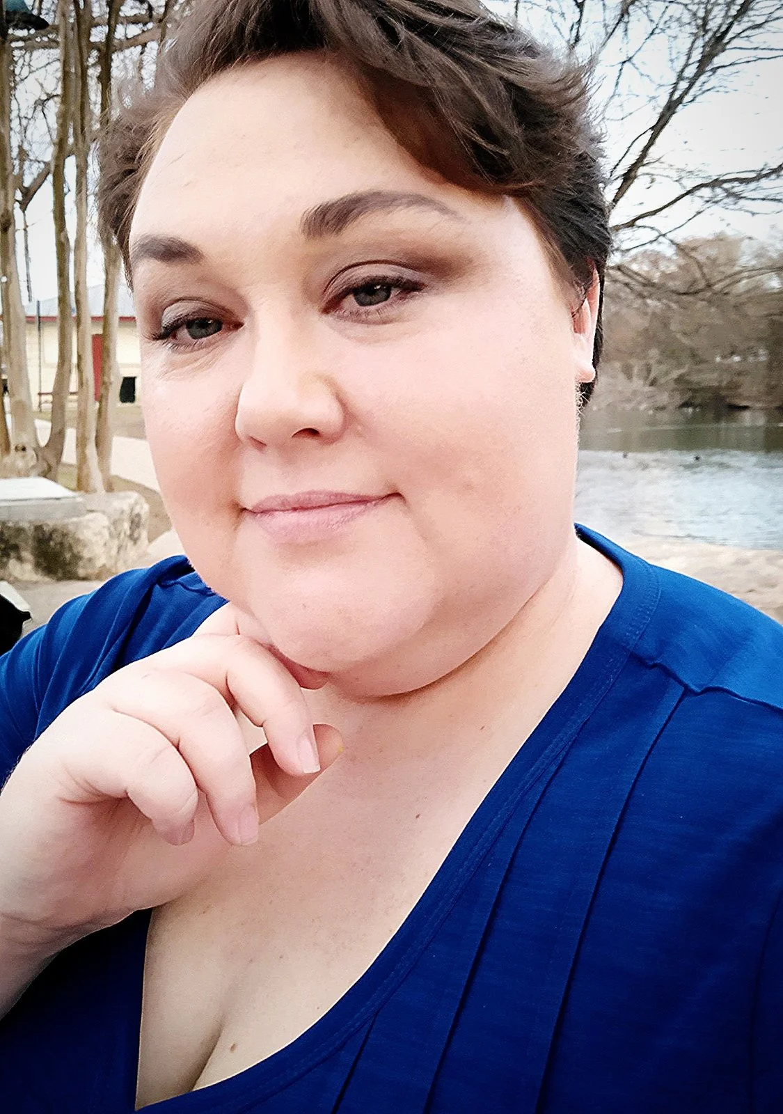 A woman with short brunette hair, wearing a blue top, is outdoors near a body of water with trees and a building in the background.