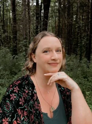 A woman with light brown hair smiling, sitting outdoors in a forest, wearing a black floral top and a necklace.