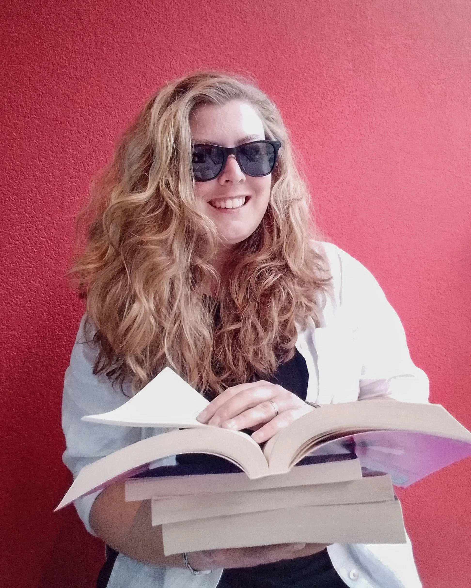 A woman with curly blonde hair wearing sunglasses, smiling, sitting in front of a red textured wall, reading a large open book.