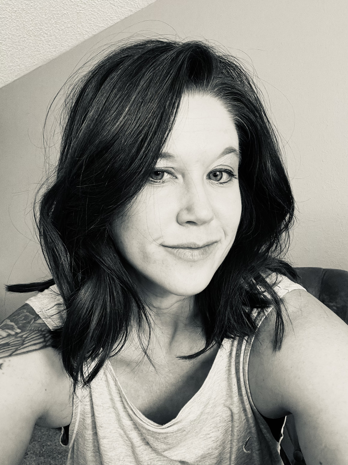 A black and white photograph of a woman with shoulder-length, wavy dark hair, smiling slightly, wearing a sleeveless top, sitting on a chair against a plain wall.