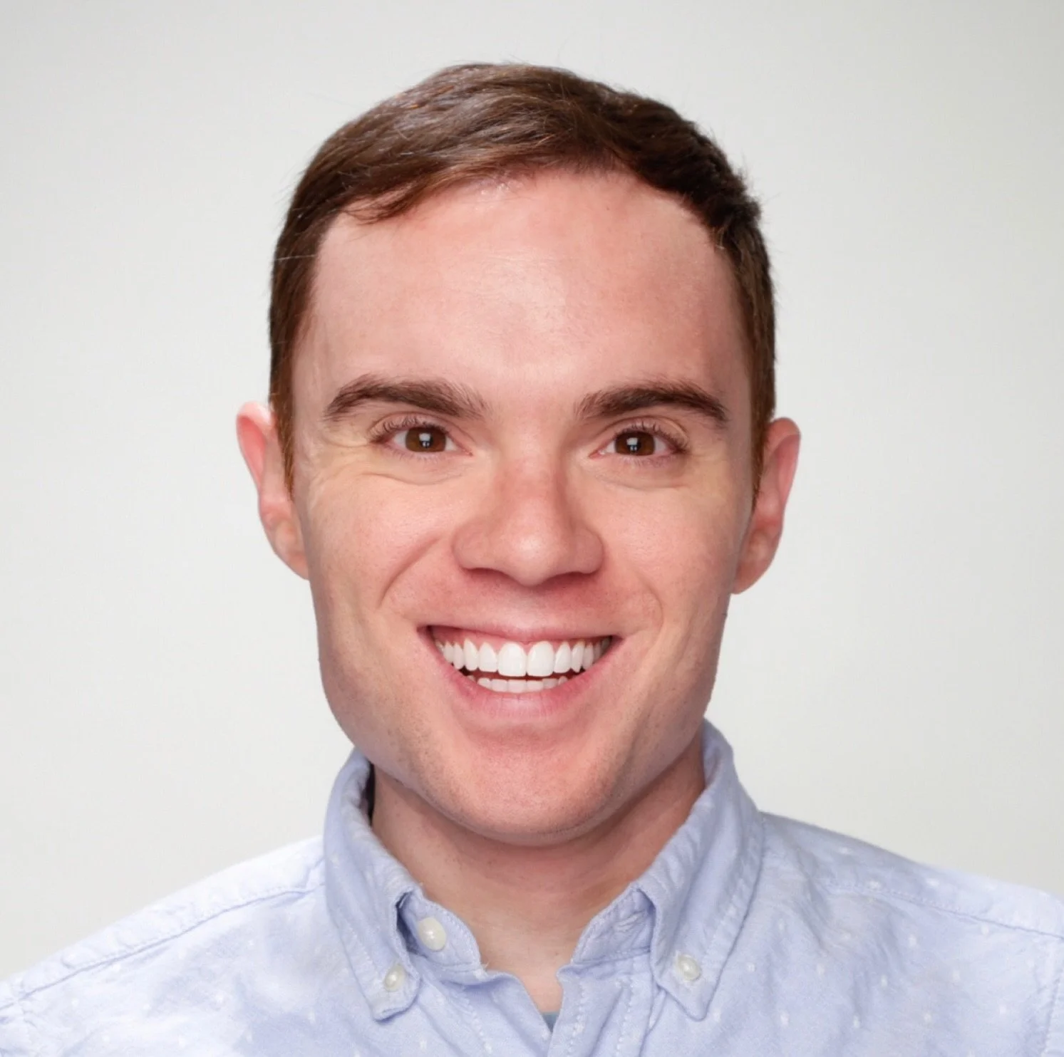 Smiling young man with brown hair, wearing a light blue button-up shirt, against a plain white background.