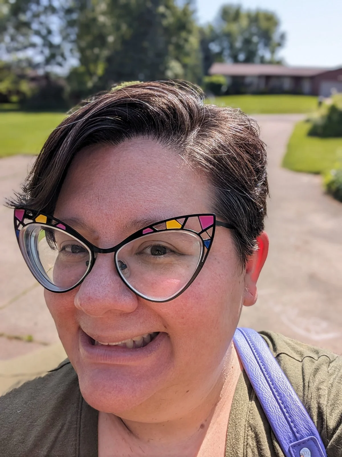 A smiling woman with short dark hair and unique cat-shaped glasses taking a selfie outdoors on a sunny day, with a green lawn and trees in the background.