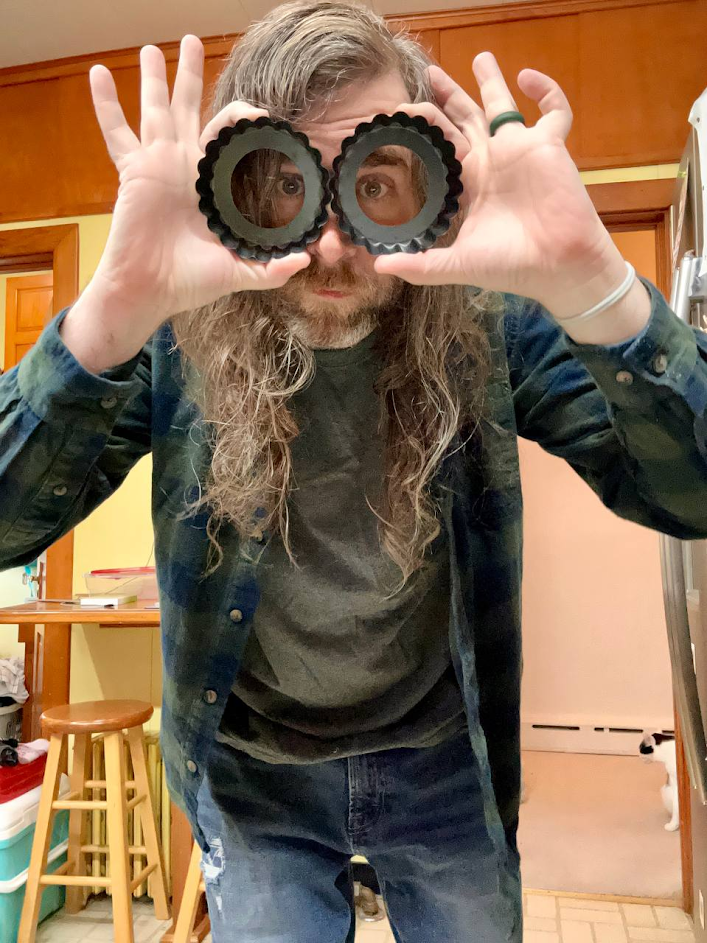 A man with long hair and a beard holding two circular objects in front of his eyes, looking through them, in a kitchen with wooden cabinets.