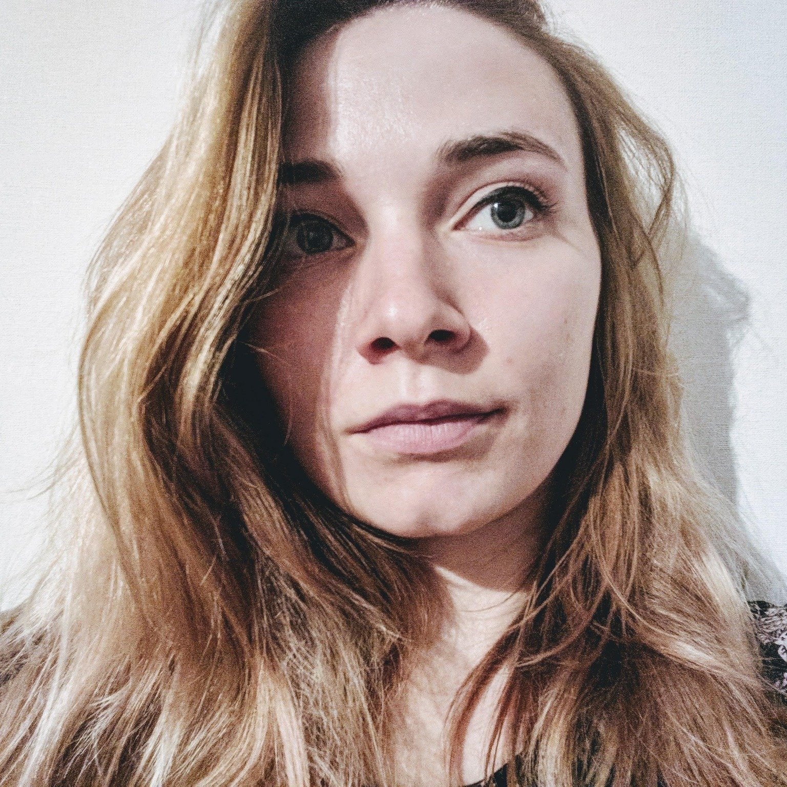 Close-up photo of a young woman with long, wavy, light brown hair and light blue eyes, looking at the camera against a plain white background.