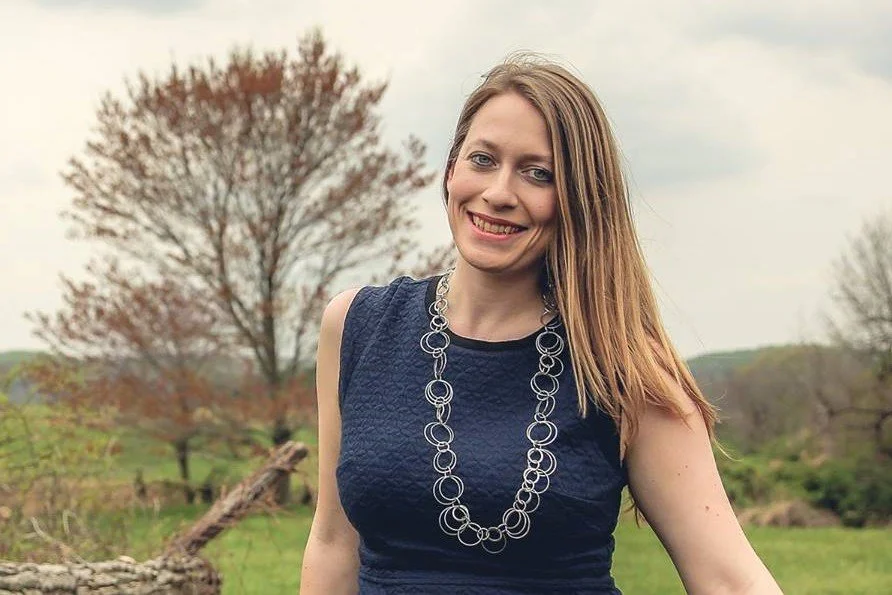 A woman with long blonde hair smiling outdoors on a cloudy day, wearing a sleeveless navy blue dress and a silver circular link necklace, standing in a grassy area with trees in the background.