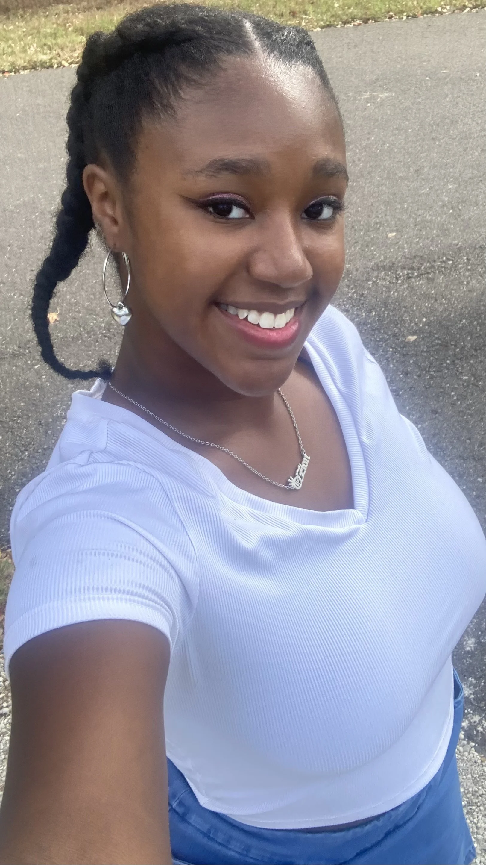 A young woman taking a selfie outdoors on a paved road with grass in the background. She has dark braided hair, is smiling, and is wearing hoop earrings, a necklace, a white ribbed shirt, and blue jeans.