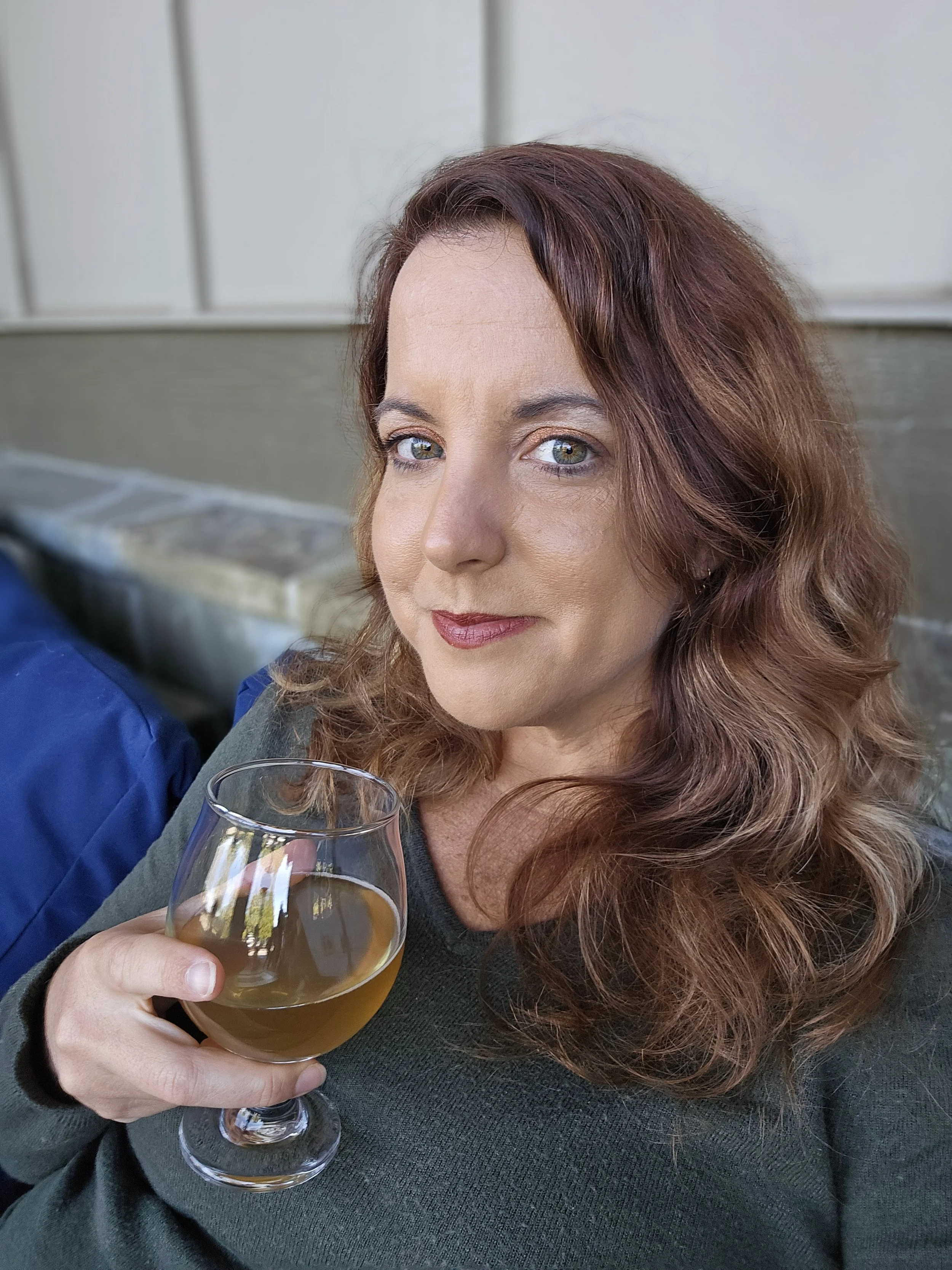 A woman with wavy red hair and blue eyes is holding a glass of beer and smiling slightly. She is wearing a dark green top and is seated outdoors, with a light-colored wall in the background.
