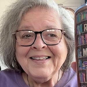 A smiling elderly woman with glasses and gray hair taking a selfie indoors.
