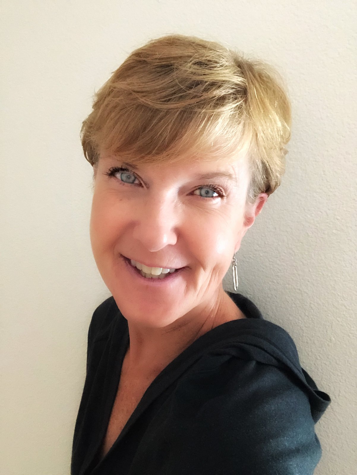 A woman with short blonde hair, blue eyes, wearing a black top, smiling, and standing against a light-colored wall.