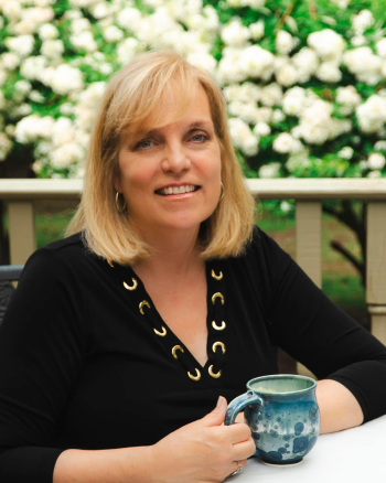 A woman with blonde hair, wearing a black top, holding a blue ceramic mug, sitting outdoors with white flowers in the background.