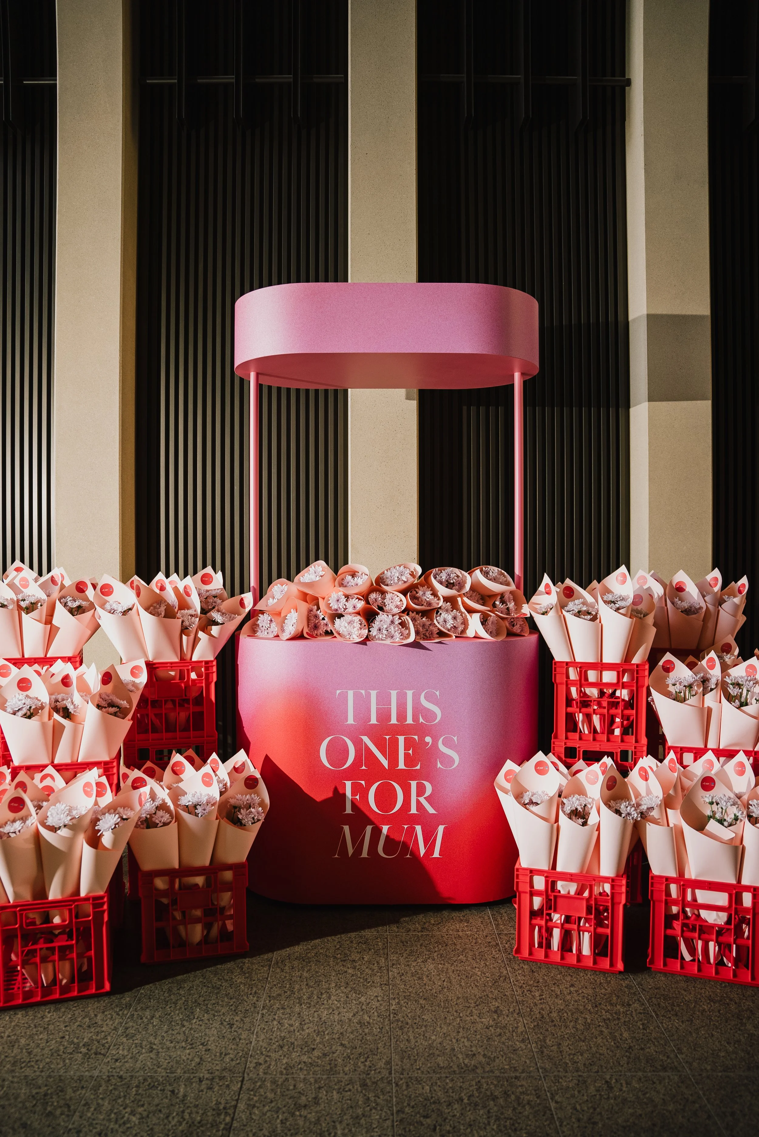A display with a pink circular stand and numerous small bouquets of flowers wrapped in pink paper, arranged in red crates. The stand has a sign that reads "This One's For Mum."