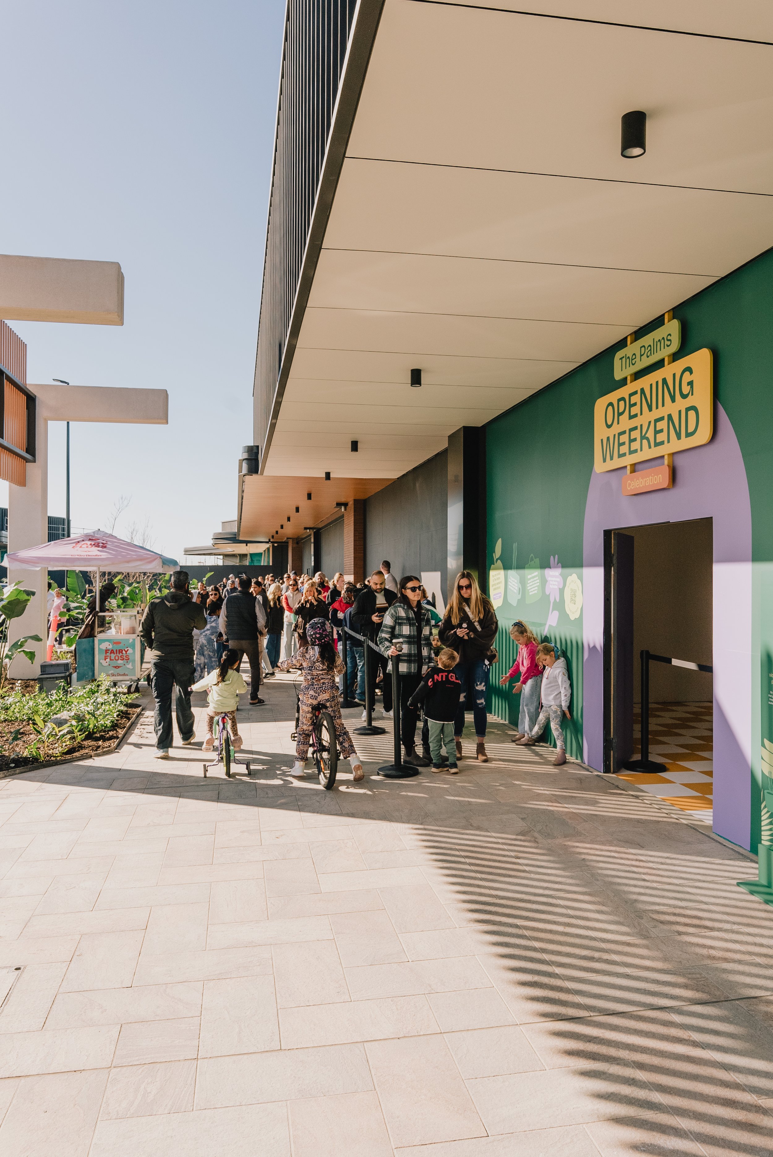 People standing and walking in line outside a building with a sign that reads 'The Palms Opening Weekend Celebration,' with some children and bicycles nearby, under clear skies.