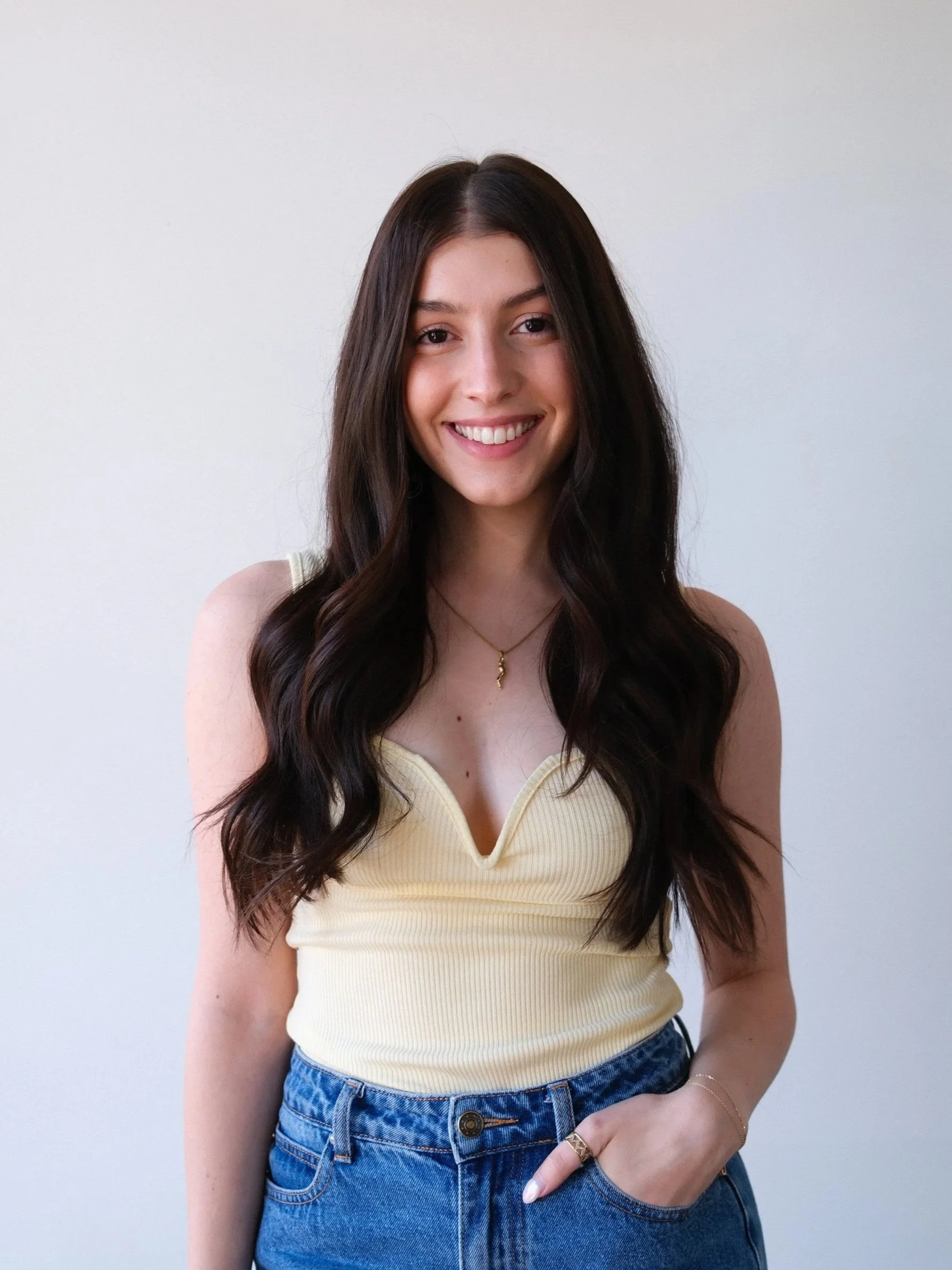 A woman with long dark hair smiling, wearing a yellow sleeveless top and blue jeans, standing against a plain white wall.