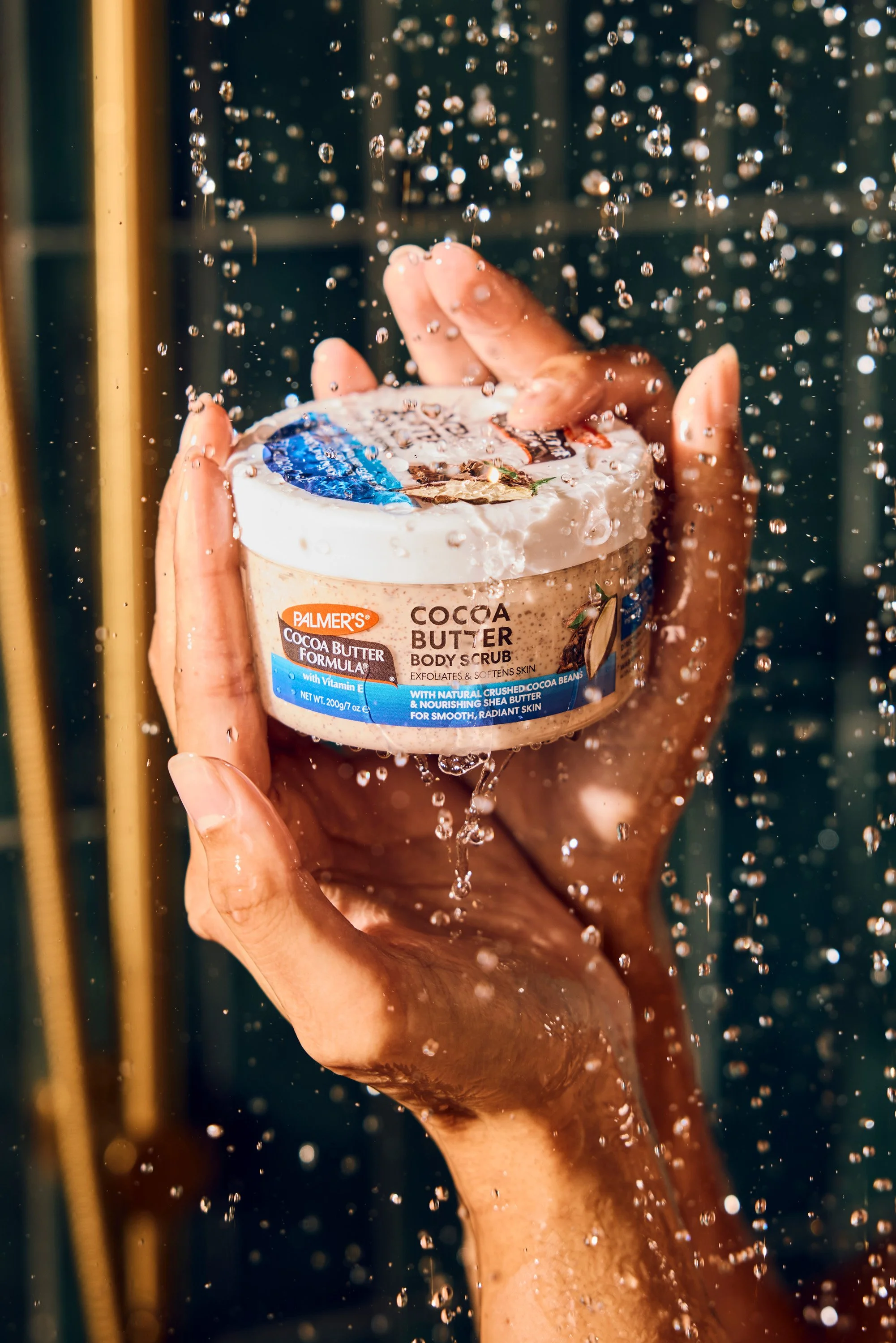 A person holding a container of Palmer's Cocoa Butter Body Scrub under running water.