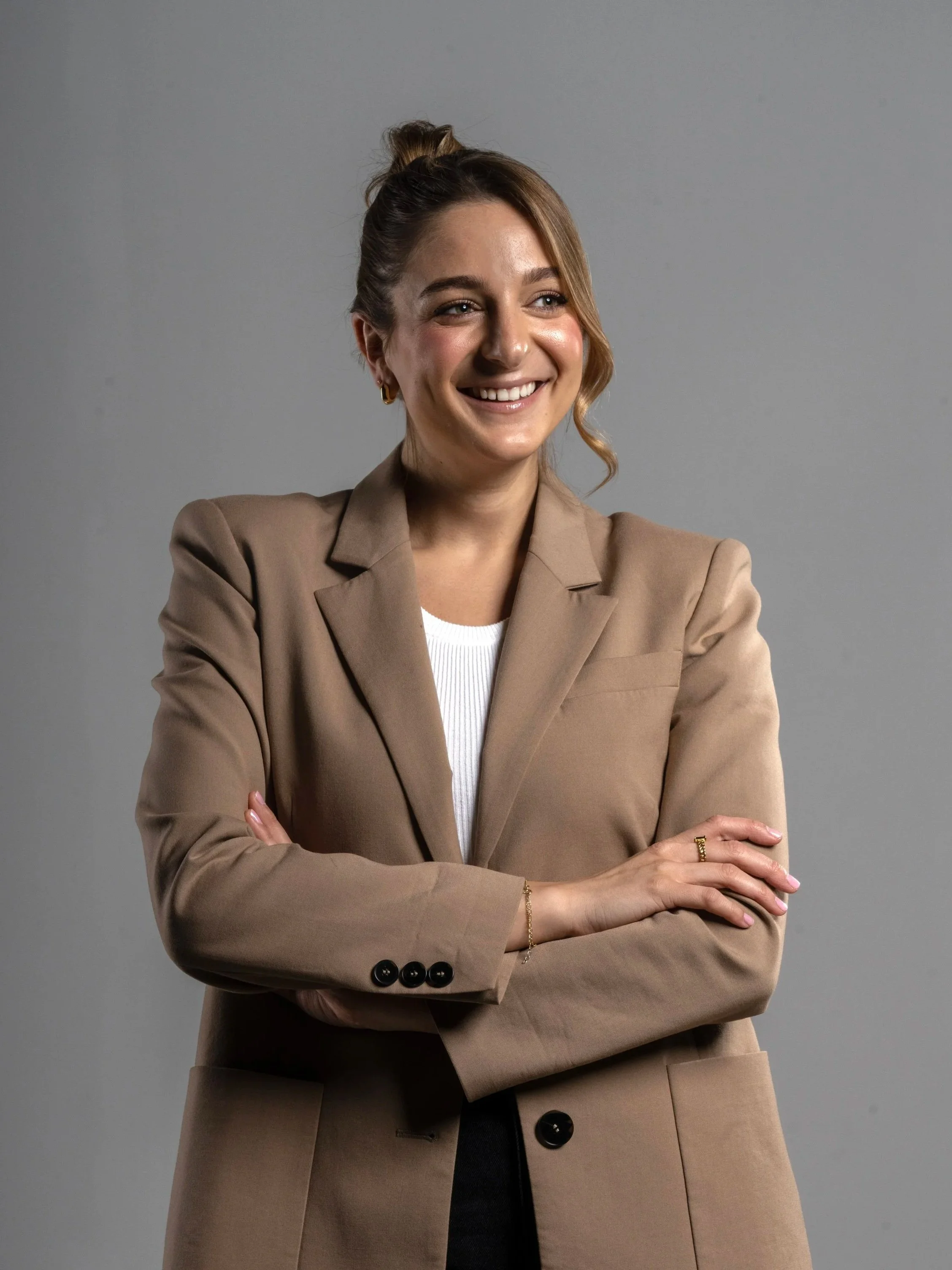 A woman with light brown hair styled in a high bun, smiling confidently with arms crossed, dressed in a beige blazer over a white top, against a plain gray background.