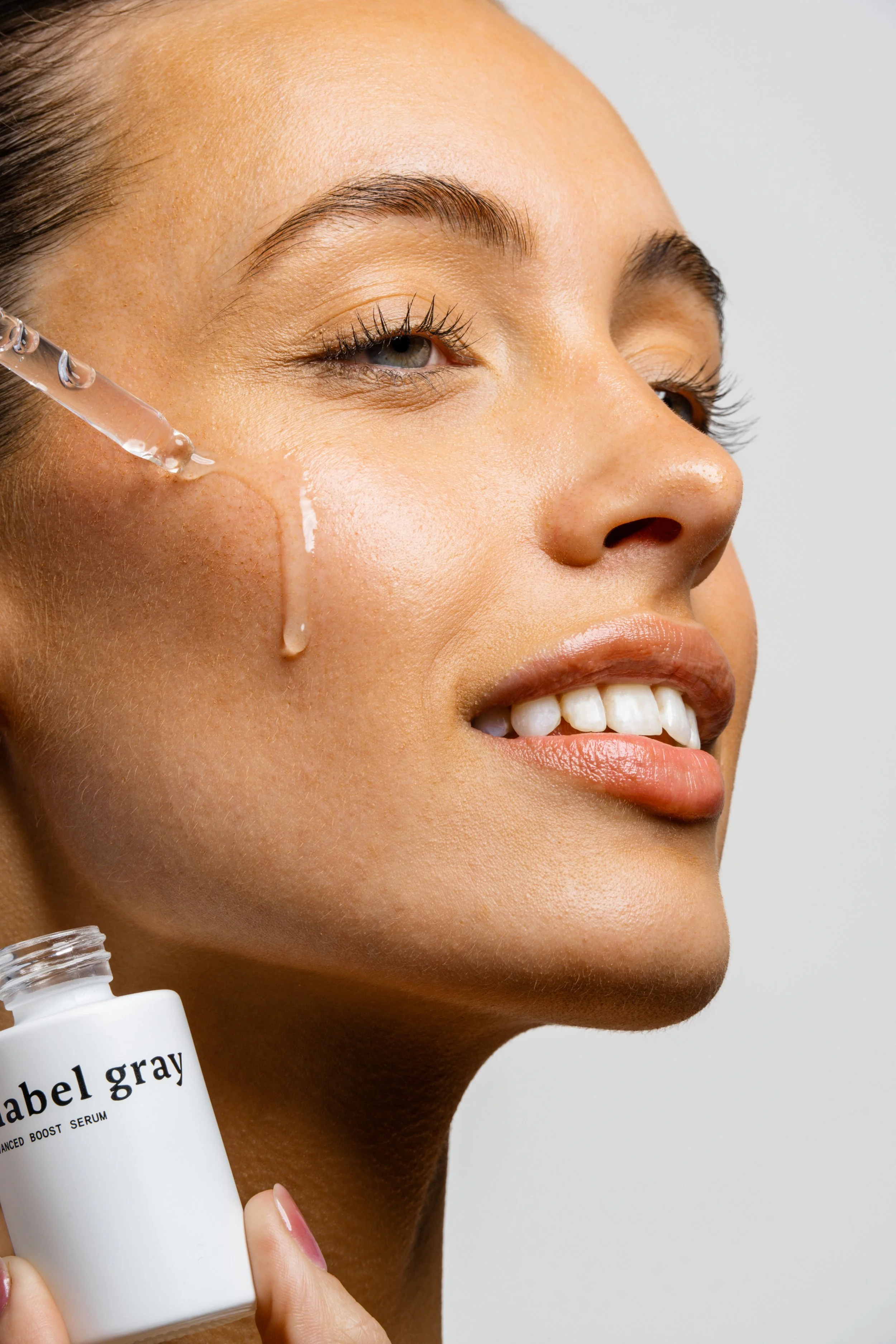 A close-up of a woman with smooth skin holding a white bottle of serums labeled 'angel gray' near her face. She is smiling softly, with a dropper applying serum to her cheek.