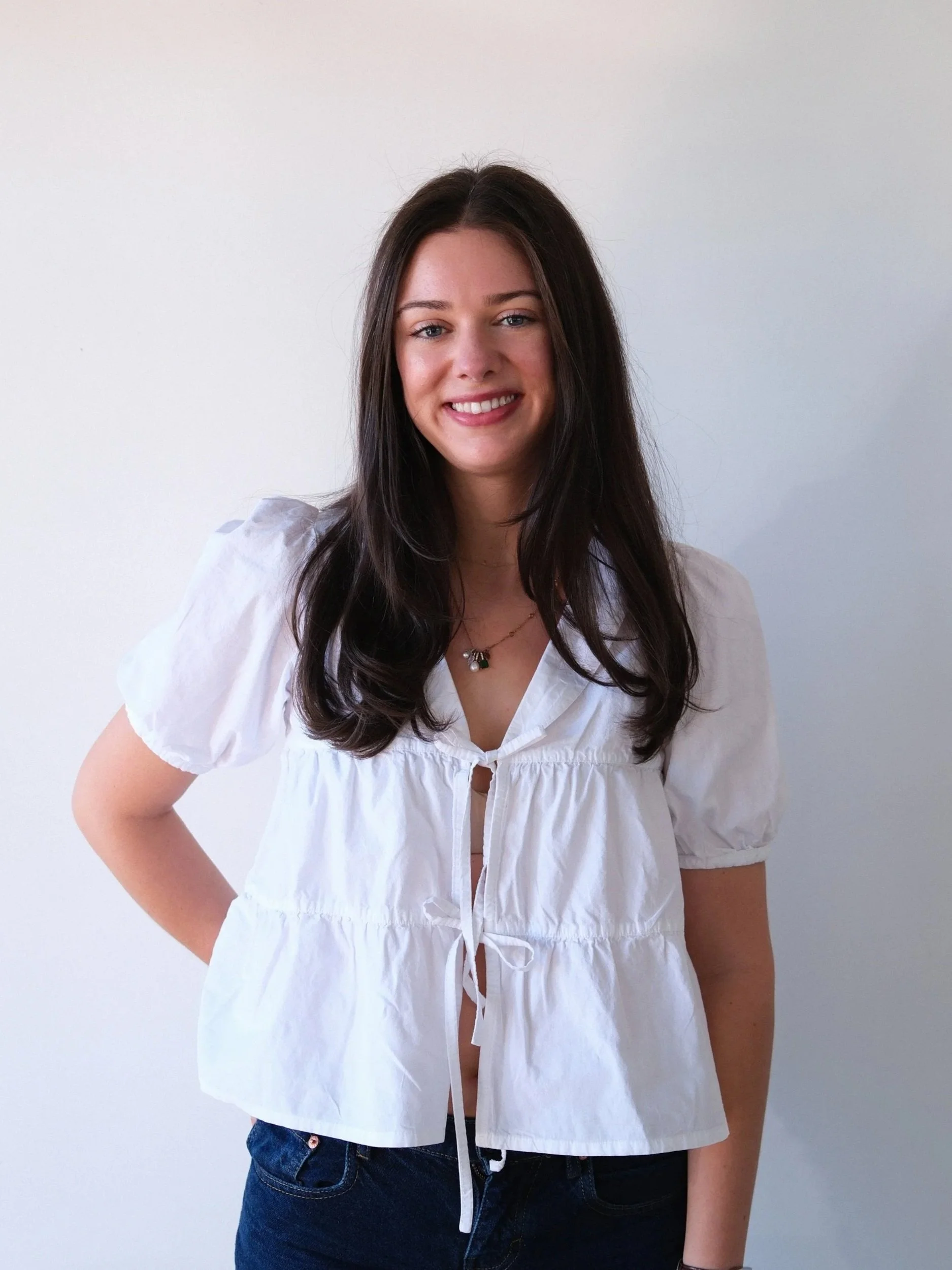 A woman with long dark hair wearing a white blouse and jeans stands against a plain white wall.