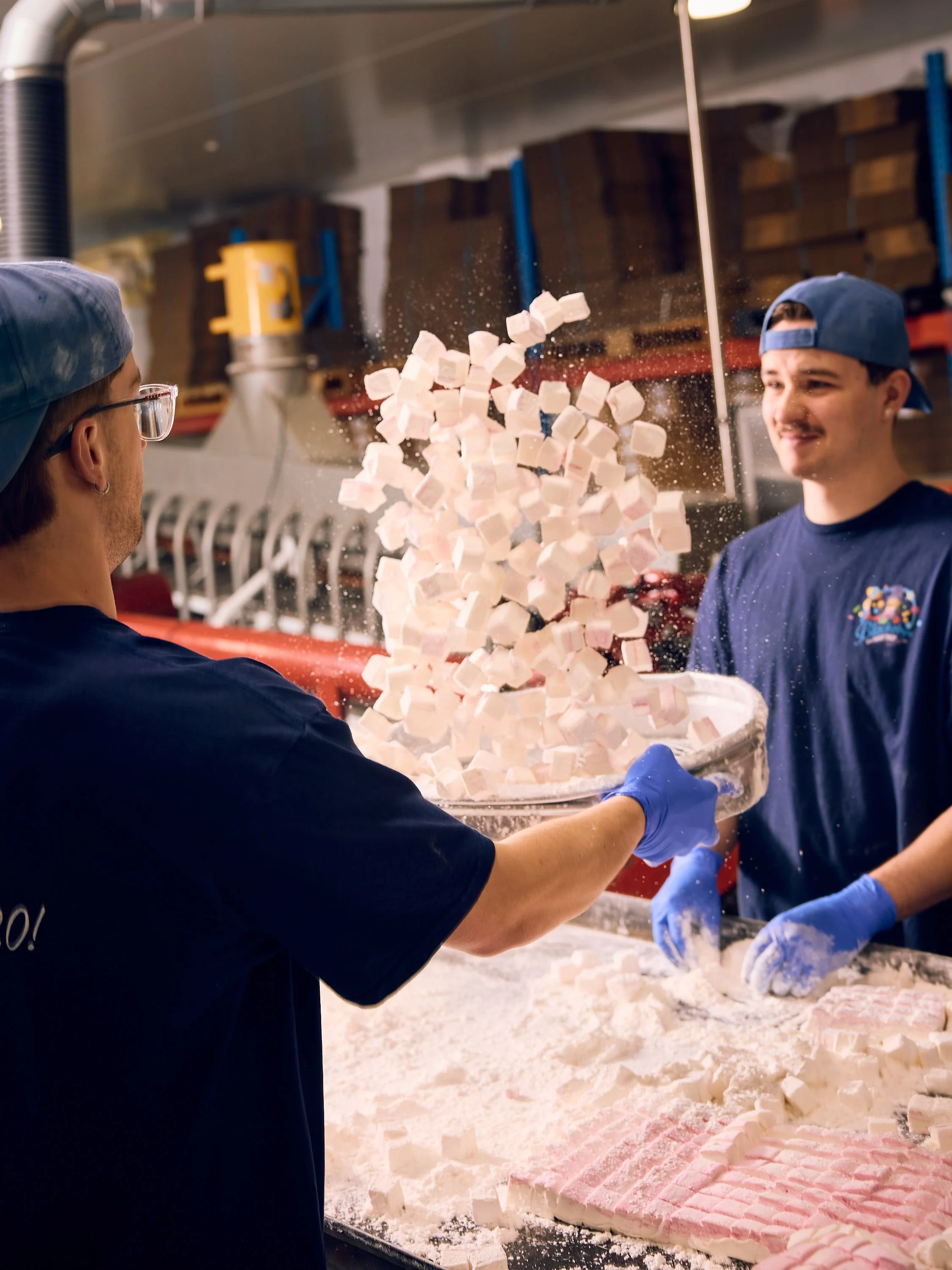 Two workers in a food manufacturing facility are handling a large tray of small, cubed, pink and white food products, possibly marshmallows or a similar item, with one worker wearing glasses and a blue hat, and the other with a backwards blue cap, while a large pizza-like object made of the same cubed food is on the table in front of them.